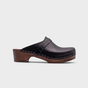 Malmö-style clogs designed by Sandgrens in classic black leather. Featuring a smooth, slip-on design with a supportive strap for stability. The sturdy wooden sole and hand-stapled construction ensure durability and all-day comfort, making them a stylish and functional footwear choice.