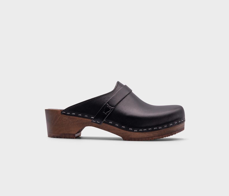 Malmö-style clogs designed by Sandgrens in classic black leather. Featuring a smooth, slip-on design with a supportive strap for stability. The sturdy wooden sole and hand-stapled construction ensure durability and all-day comfort, making them a stylish and functional footwear choice.