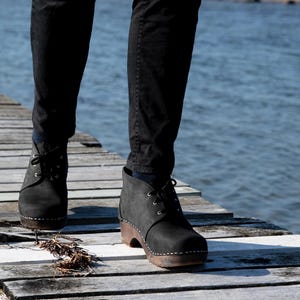 Könnte beinhalten: Schwarze Lederstiefel mit Schnürsenkeln und einer Holzplattformsohle. Die Stiefel werden von einer Person getragen, die auf einem Holzsteg geht.