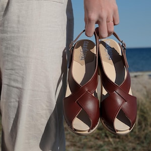 Könnte beinhalten: Ein Paar braune Ledersandalen mit einem Kreuzriemen-Design. Die Sandalen haben eine Holzsohle und einen Slingback-Riemen.