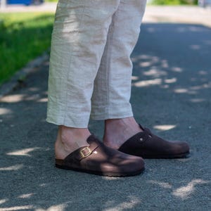Puede incluir: Un par de zuecos de cuero marrón con una hebilla. Los zuecos los lleva una persona que lleva pantalones beige.