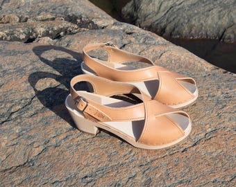 May include: A pair of light brown leather heeled sandals with a criss-cross strap design. The sandals are on a rocky surface.
