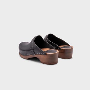 Malmö-style clogs designed by Sandgrens in classic black leather. Featuring a smooth, slip-on design with a supportive strap for stability. The sturdy wooden sole and hand-stapled construction ensure durability and all-day comfort, making them a stylish and functional footwear choice.