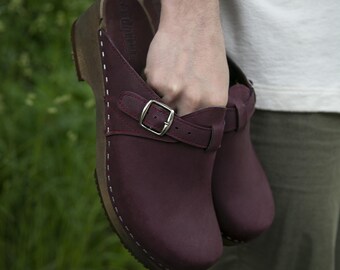 May include: A pair of burgundy leather clogs with a buckle strap. The clogs have a wooden base and are stitched around the edges.
