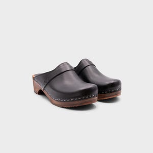 Malmö-style clogs designed by Sandgrens in classic black leather. Featuring a smooth, slip-on design with a supportive strap for stability. The sturdy wooden sole and hand-stapled construction ensure durability and all-day comfort, making them a stylish and functional footwear choice.