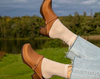 Puede incluir: Un par de zuecos de cuero marrón con tacón de madera. Los zuecos se llevan con calcetines y pantalones vaqueros.