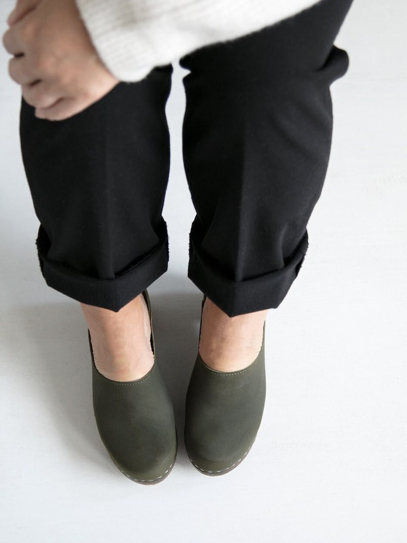 woman wearing black dress pants, a white knitted sweater and high heeled closed-back clogs in dark green leather on a dark wooden base