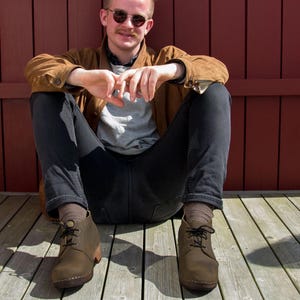 Könnte beinhalten: Eine Person trägt eine braune Wildlederjacke, eine dunkelblaue Jeans und braune Lederstiefel mit Schnallen. Die Person sitzt auf einem Holzdeck mit einer roten Holzwand dahinter.