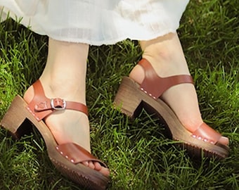 May include: A pair of brown leather heeled sandals with a buckle closure. The sandals are worn by a person standing on green grass.