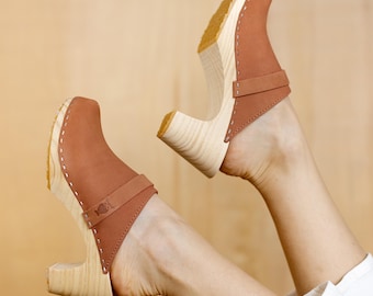Puede incluir: Un par de zuecos de cuero marrón con tacones de madera. Los zuecos tienen una correa en la parte superior del pie y se llevan puestos.