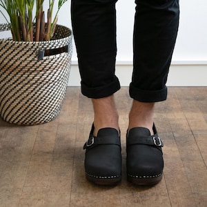 Puede incluir: Zuecos de cuero negro con una correa de hebilla. Los zuecos tienen una suela de madera y los lleva una persona con pantalones negros remangados.
