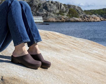 May include: A pair of brown leather clogs with a wooden heel. The clogs are worn by a person sitting on a rocky surface near a body of water.