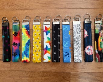 May include: Ten fabric wristlet keychains with metal hardware and split rings. Patterns include black with paw prints, rainbow tie-dye, yellow with bees, colorful stars, black with Om symbols, blue with anchors, white with nurse caps, baseball logos, and beer bottles.