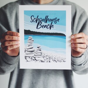 May include: A white print with blue and green accents featuring the text "Schoolhouse Beach" and "Washington Island". The print depicts a beach scene with stacked rocks and a blue body of water.