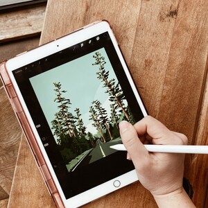 May include: A digital illustration of a road through a forest on a tablet screen. A hand is holding a stylus and drawing on the screen.