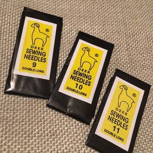 May include: Three black packets of Deer Sewing Needles, sizes 9, 10, and 11, are displayed. Each packet has a yellow label with a deer logo and the text "DOUBLE LONG". The packets are arranged on a textured, neutral-toned surface.