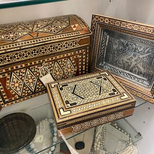 May include: Three wooden boxes with intricate inlaid designs. The boxes are made of various woods and have a dark brown finish. The largest box is a chest with a hinged lid. The middle box is a rectangular box with a lid. The smallest box is a square frame with a silver-colored metal insert.