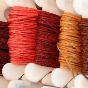 Puede incluir: Seis carretes de hilo de bordar en varios tonos de rojo, marrón y beige. El hilo está enrollado alrededor de carretes de plástico blanco con una cubierta de plástico transparente.