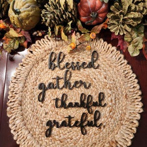 Op de afbeelding: Een ronde placemat van jute met de woorden "blessed, gather, thankful, grateful" in zwart script. De placemat is omgeven door herfstdecoraties, waaronder pompoenen, bladeren en dennenappels.
