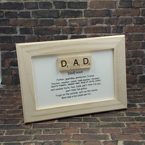 May include: A framed print with a definition of the word "dad" spelled out with wooden letter tiles. The definition reads: "Father, guardian, protector, friend. Teacher, mentor, coach, task master, listener. Sports fanatic, mower man, king of corny jokes and useless facts, taxi, bank, part-time fun guy, part-time fun police. Tough on the outside, soft on the inside. Best dad a kid could ask for..."