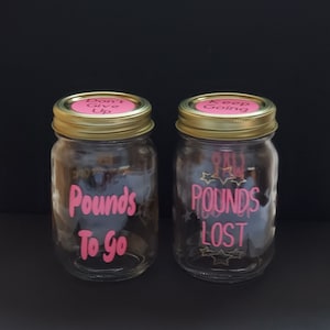 May include: Two glass jars with gold lids and pink labels. The jar on the left has the text "Don't Give Up" on the lid and "Pounds To Go" on the jar. The jar on the right has the text "Keep Going" on the lid and "Pounds Lost" on the jar.