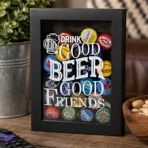 May include: A black rectangular shadow box frame containing a collection of colorful metal beer bottle caps. The glass front features white text that reads "DRINK GOOD BEER WITH GOOD FRIENDS" along with a graphic of a beer mug icon.