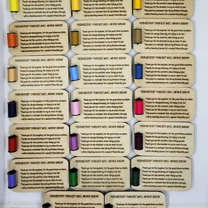 Puede incluir: Veinticuatro placas de madera con carretes de hilo de colores unidos. Cada placa presenta un poema sobre la amistad y el texto "Friendship Threads Will Never Break".