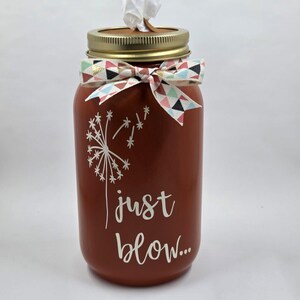 Op de afbeelding: Een terracotta-kleurige pot met een gouden metalen deksel en een wit doekje dat uit de bovenkant steekt. De pot is voorzien van een witte paardenbloemafbeelding en de tekst "just blow..." in wit schrift. Een lint met geometrisch patroon.