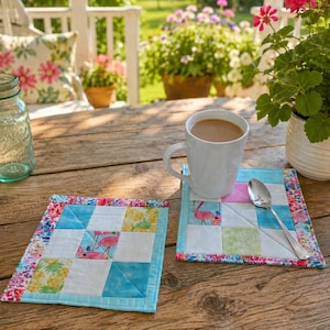 May include: Two square quilted coasters with a flamingo and pineapple pattern, a white mug with coffee, and a spoon rest on a wooden table. The coasters have a light blue border and a colorful floral patterned edge.