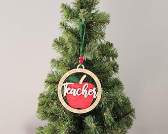 Può includere: Un ornamento natalizio in legno con un design a mela rossa e la parola "Teacher" in corsivo bianco. L'ornamento è circondato da un cerchio con parole come "Inspiring" e "Caring". È appeso a un nastro verde su un piccolo albero di Natale.