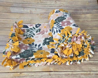 May include: A fleece blanket featuring a floral pattern with mustard yellow, dark green, and dusty pink flowers and leaves on a white background. The edges of the blanket are finished with a knotted fringe made from strips of the same fleece fabric.