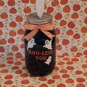Op de afbeelding: Een zwarte glazen pot als tissuehouder met witte spookillustraties en de oranje tekst BOO-LESS YOU. De pot heeft een zilveren metalen deksel met een witte tissue die eruit steekt en een oranje met witte gingham strik aan de hals.