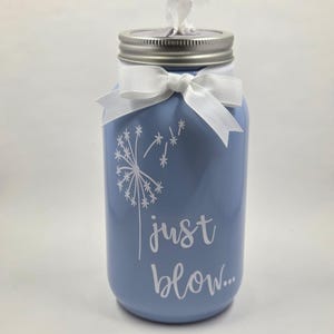Op de afbeelding: Een lichtblauwe cilindervormige tissuehouder met een metalen schroefdeksel. De voorzijde is voorzien van een witte paardenbloemafbeelding en de tekst "just blow..." in een sierlijk lettertype. Een witte strik zit om de hals, met een witte tissue.