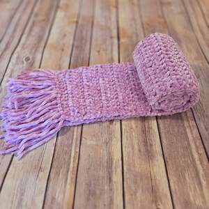 May include: A lavender-colored scarf crafted from thick, plush chenille yarn. The scarf features a soft, velvety texture with a rolled end and a fringe of long, matching yarn strands at the opposite end, creating a cozy and tactile appearance.