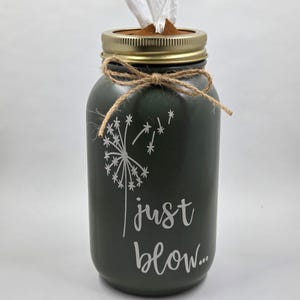 Op de afbeelding: Een donkergroene, cilindrische tissuehouder gemaakt van een glazen pot met een goudkleurig metalen deksel. De pot is voorzien van een witte paardenbloemafbeelding en de tekst "just blow..." in wit schrift. Een dun jute strikje zit om de hals.