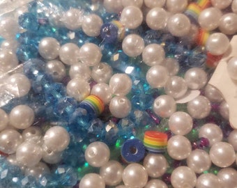 May include: A close-up image of a variety of beads, including white pearl beads, blue crystal beads, and rainbow-colored beads. The beads are arranged in a random pattern and are suitable for jewelry making.