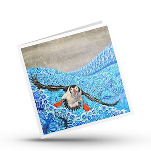May include: A square greeting card featuring a colorful painting of a puffin in flight over stylized blue ocean waves. The bird has black and white feathers, a bright orange beak holding a small fish, and orange webbed feet against a textured sky.