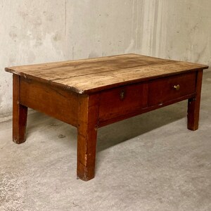 Peut inclure: Une table basse en bois rustique avec deux tiroirs et des poignées en laiton. La table a un plateau rectangulaire et quatre pieds.