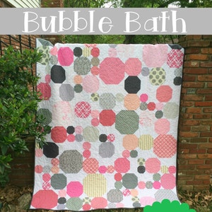 May include: A quilt pattern called "Bubble Bath" by Kelly Young. The quilt is a modern snowball design with pink, grey, and white fabric. The quilt is hanging on a brick wall with green bushes in the background.
