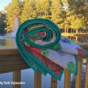 Pu&ograve; includere: Un quilt arrotolato con un motivo floreale turchese, verde, rosa e bianco. Il quilt &egrave; appoggiato su una ringhiera di legno che si affaccia su uno stagno con alberi sullo sfondo.