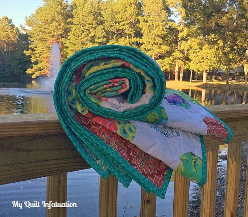 Pu&ograve; includere: Un quilt arrotolato con un motivo floreale turchese, verde, rosa e bianco. Il quilt &egrave; appoggiato su una ringhiera di legno che si affaccia su uno stagno con alberi sullo sfondo.