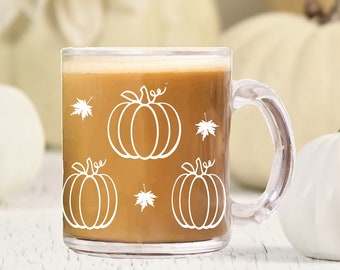 May include: A clear glass mug with a brown liquid inside. The mug has a white outline of pumpkins and leaves on the side. The mug is sitting on a white wooden surface with white pumpkins in the background.