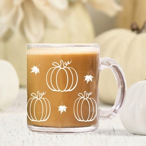 Puede incluir: Una taza de vidrio transparente con un l&iacute;quido marr&oacute;n en su interior. La taza tiene un contorno blanco de calabazas y hojas en el lateral. La taza est&aacute; sobre una superficie de madera blanca con calabazas blancas en el fondo.