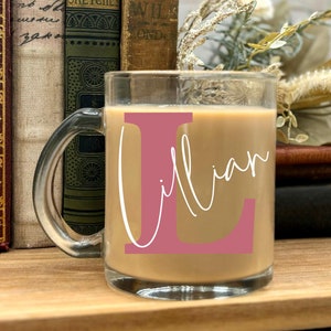 以下が含まれることがあります： 透明なガラス製のマグカップに、ピンクのLの文字と白い筆記体で「Lillian」という名前が書かれています。マグカップには薄い茶色の飲み物が注がれており、木製の表面の上に置かれています。