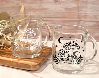 May include: Two clear glass mugs with black and white mushroom designs. The mugs feature a crescent moon and stars. One mug has white mushrooms and the other has black mushrooms.