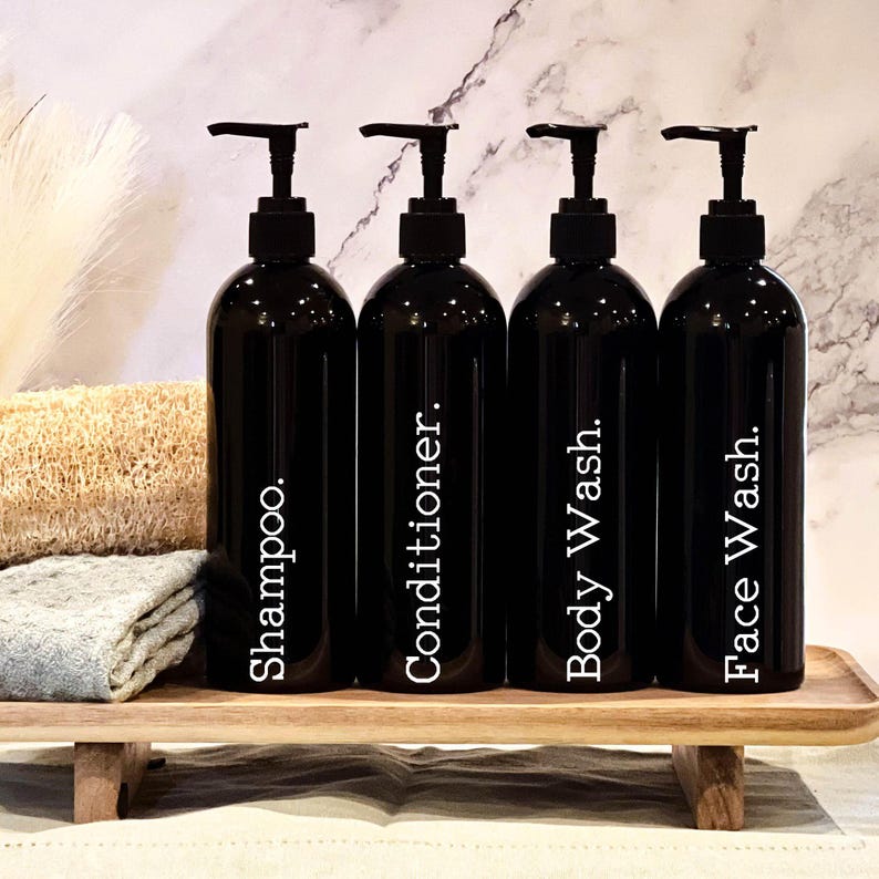 May include: Four black pump bottles with white labels on a wooden shelf. The labels read "Shampoo", "Conditioner", "Body Wash", and "Face Wash".