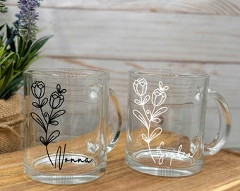 May include: Two clear glass mugs with black and white floral designs and personalized names. The mug on the left has a black floral design and the name "Nonna" written below. The mug on the right has a white floral design and the name "Sophia" written below.