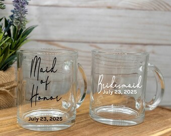 May include: Two clear glass mugs with handles. One mug says "Maid of Honor" with the date "July 23, 2025". The other mug says "Bridesmaid" with the date "July 23, 2025".