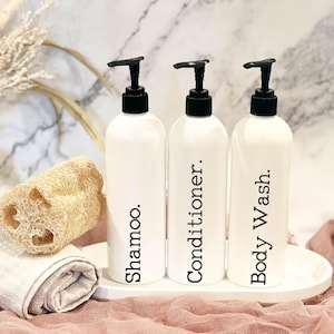 May include: Three white pump bottles with black labels and black pumps. The labels read "Shampoo", "Conditioner", and "Body Wash".