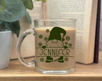 May include: A green gnome with a white beard and a red hat holding a sign that says "Jennifer". The gnome is surrounded by snowflakes. The gnome is on a clear glass mug with a brown liquid inside.